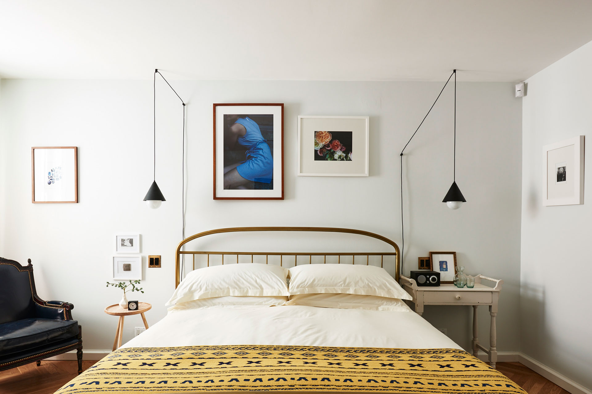 A bedroom with white walls is well decorated with a hanging light lamp. Several portraits are hanging on the wall just above the bed.