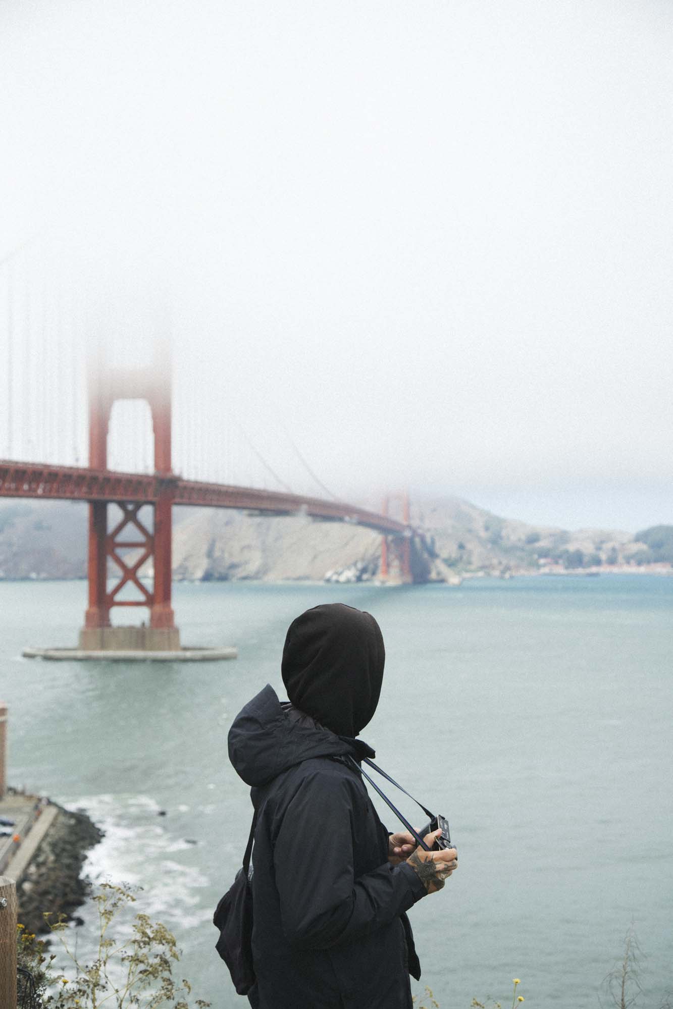 Person in black hoodie stands in front of large red bridge