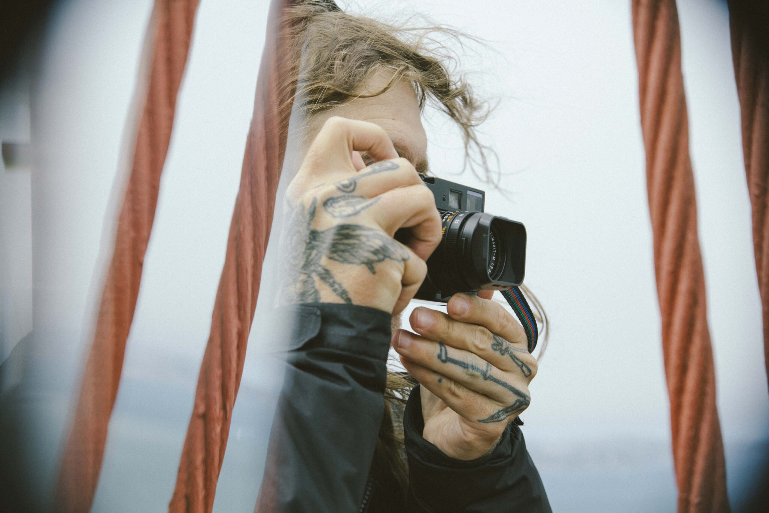 A person with tattoos on their hands is taking a photo next to the suspension bridge cables.