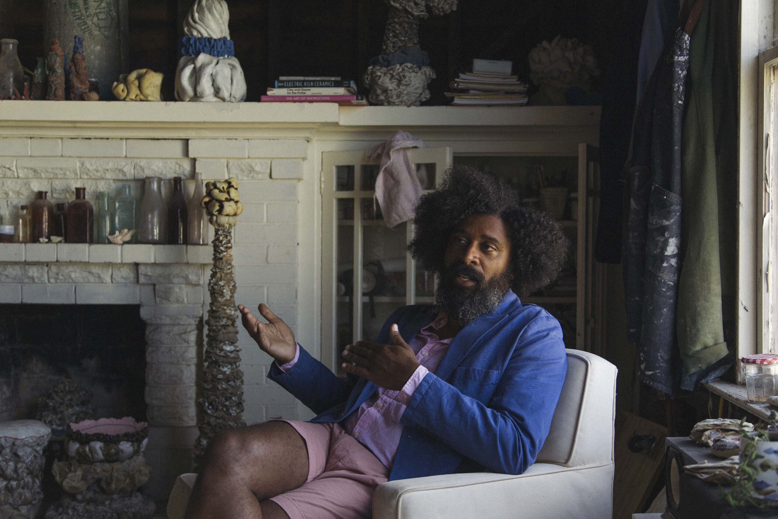 A man is seated in a chair near the fireplace, making gestures with his hands.