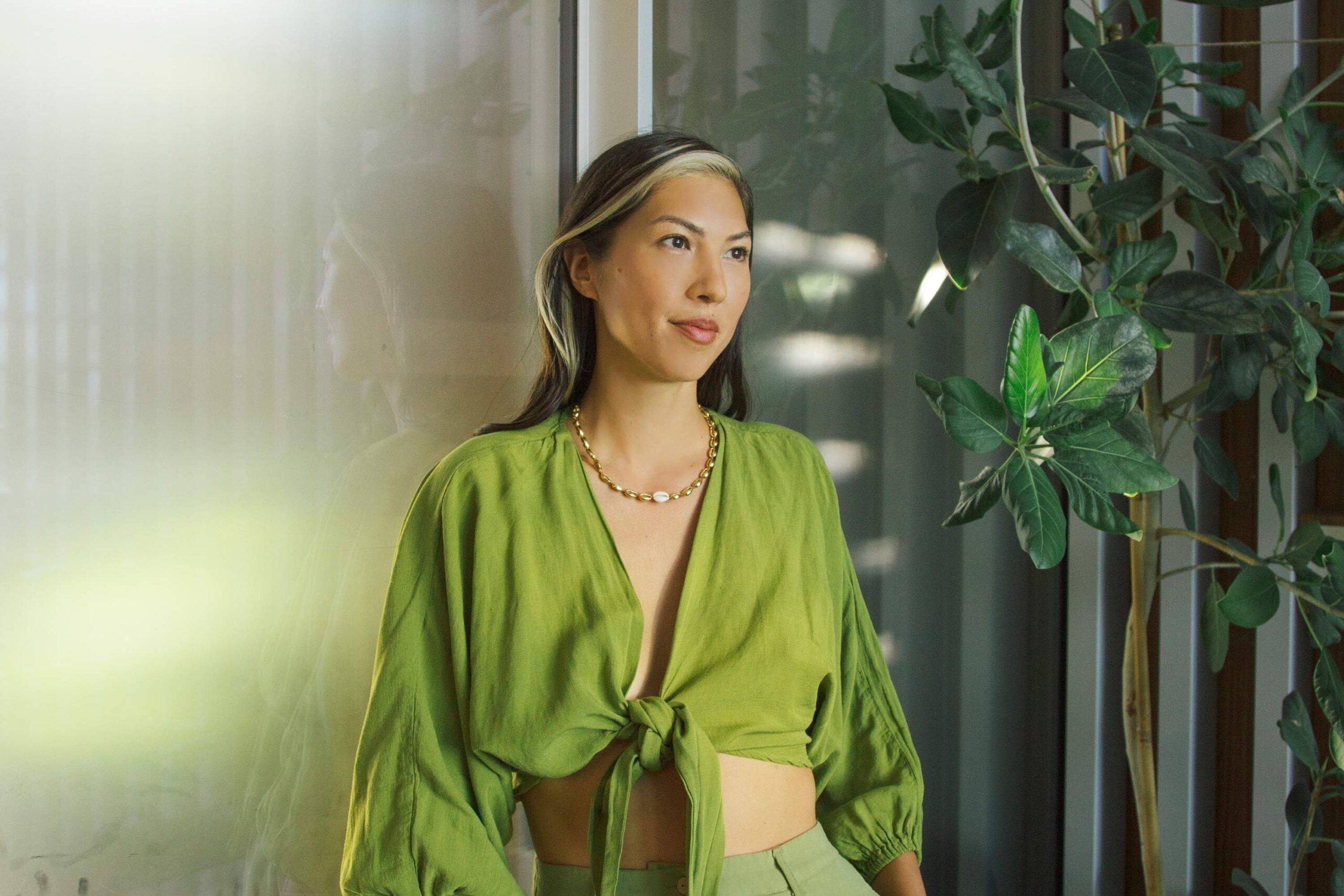 A woman wearing a green top is standing in front of a glass door and looking towards her right side.