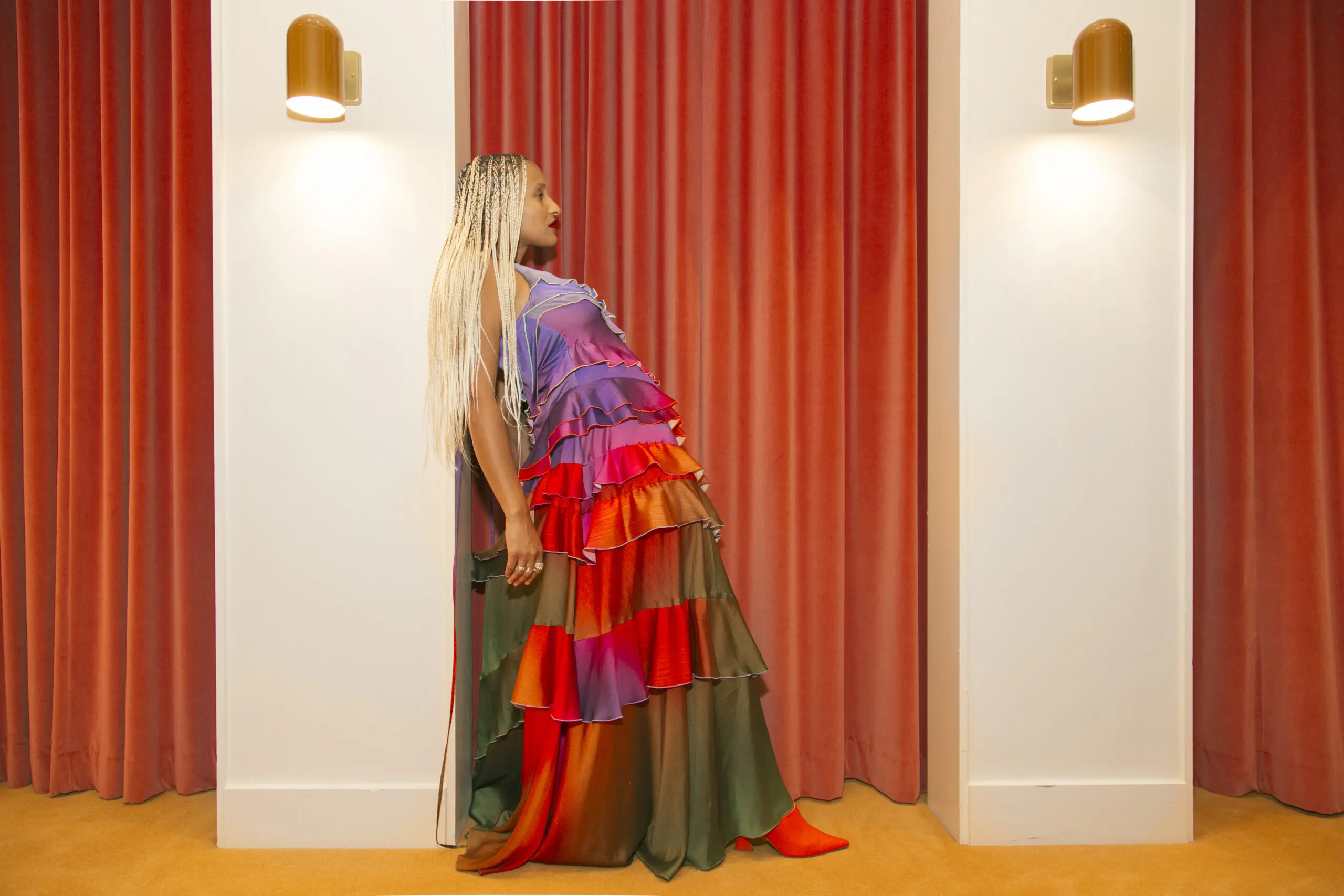 A woman wearing a colorful gown dress is leaning against a wall that is situated in front of a luxurious curtain.