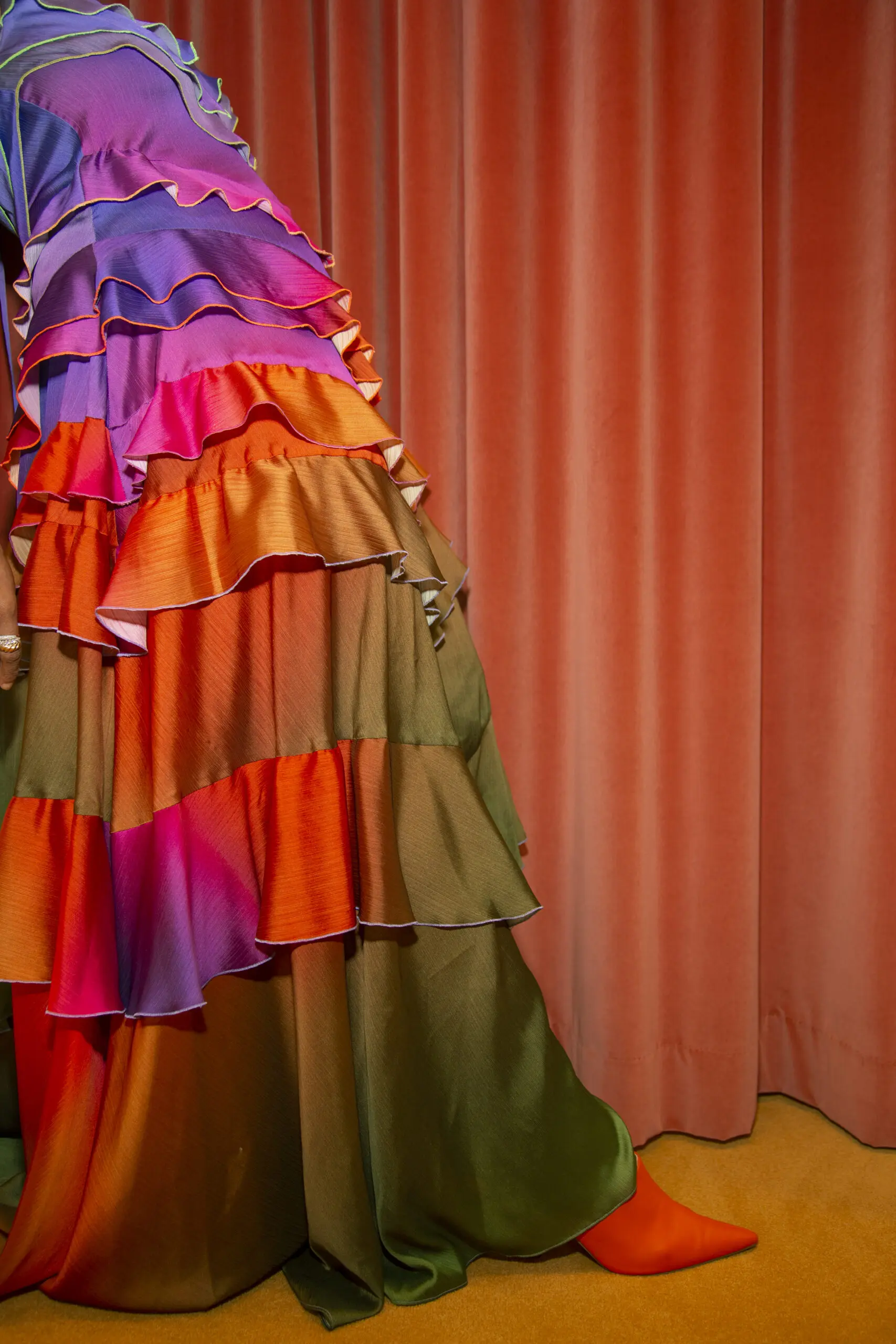 A woman is standing next to a red curtain, wearing a colorful frock dress.