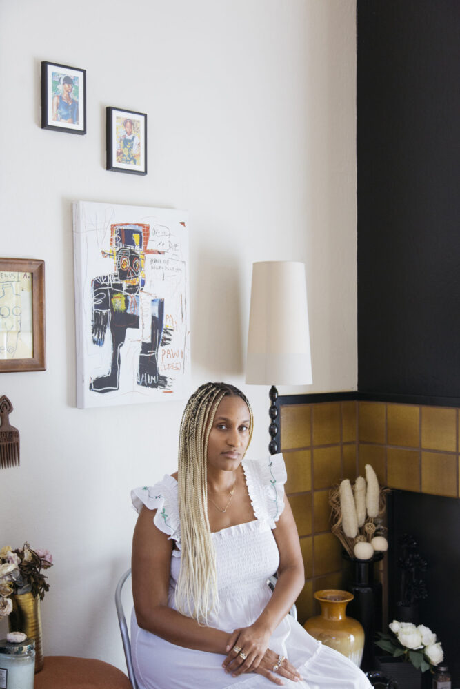 A woman wearing a white dress is sitting on a chair beside the wall, and there is a handcrafted portrait hanging on the wall.