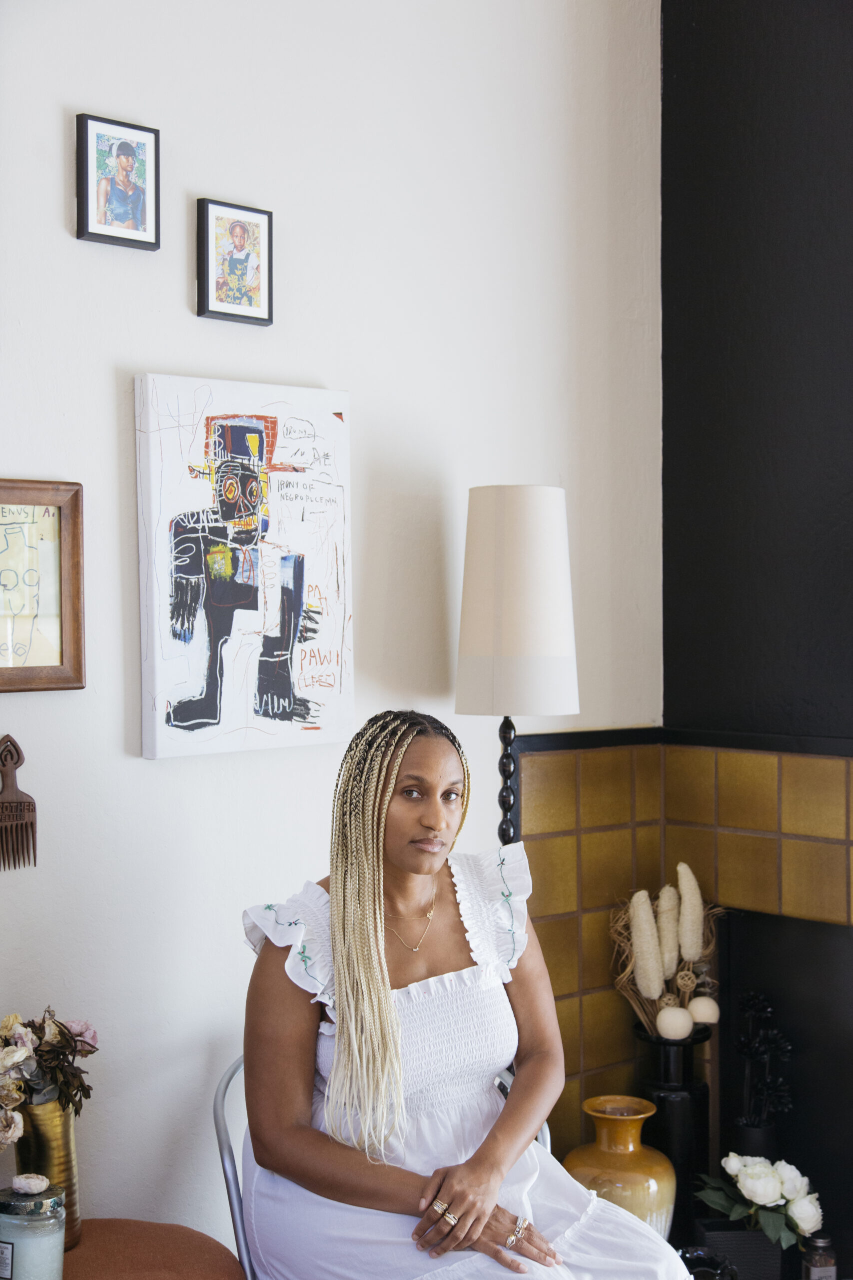 A woman wearing a white dress is sitting on a chair beside the wall, and there is a handcrafted portrait hanging on the wall.