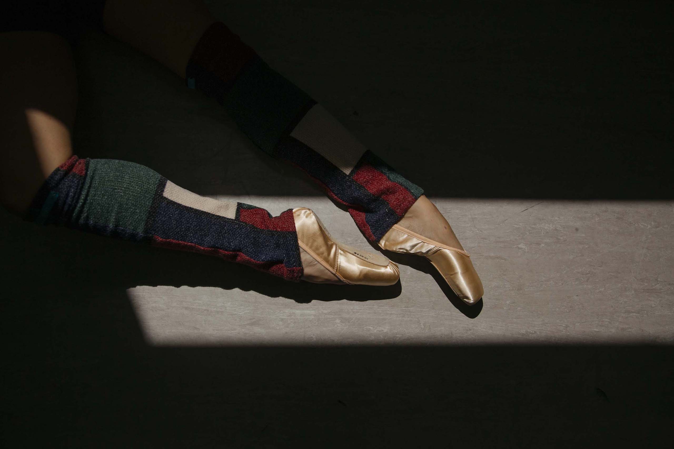 The lower legs of an individual wearing ballet shoes and colorful leg warmers.