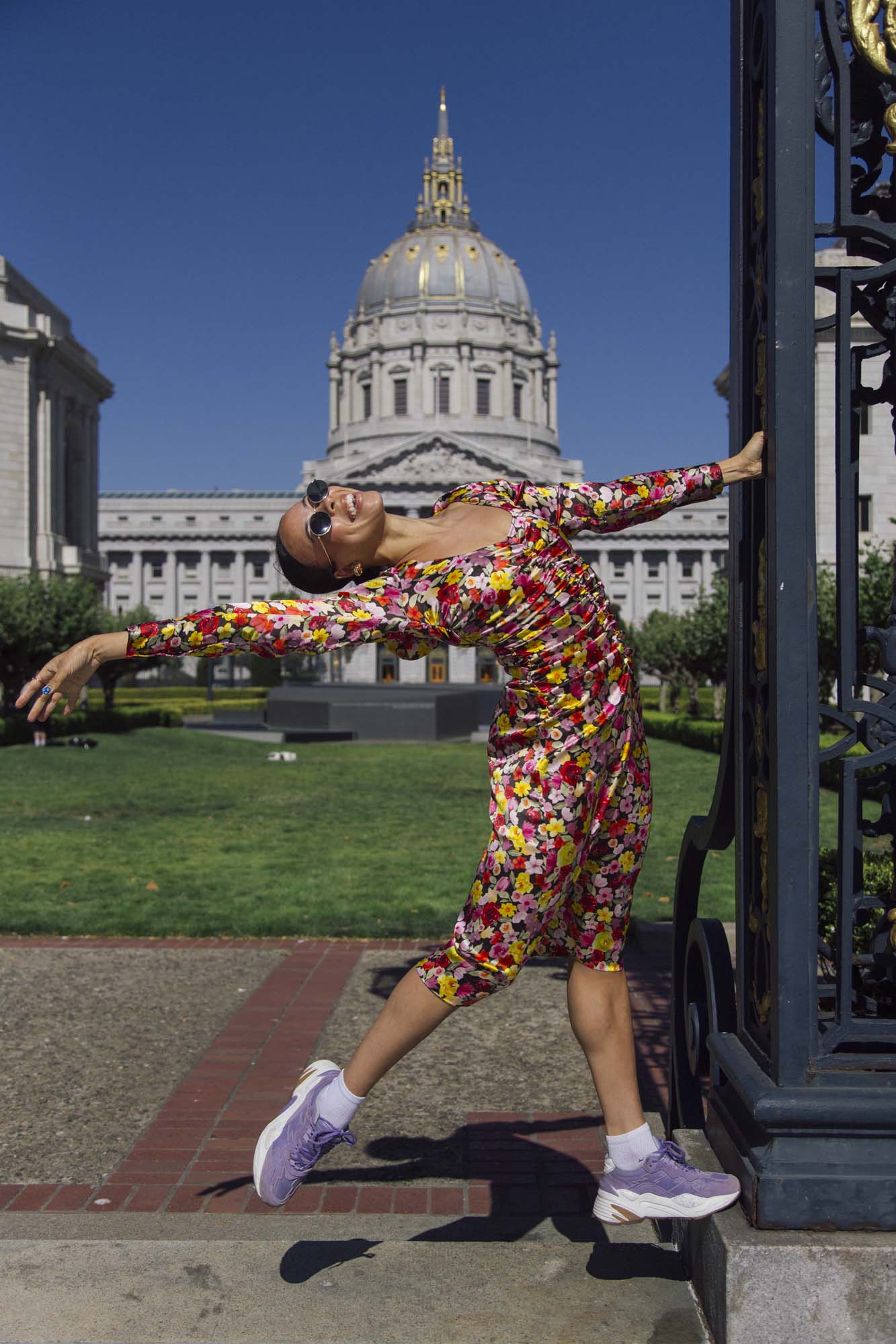 A woman dressed in a floral dress is posing for a photo while holding an iron pillar in front of a stately building.