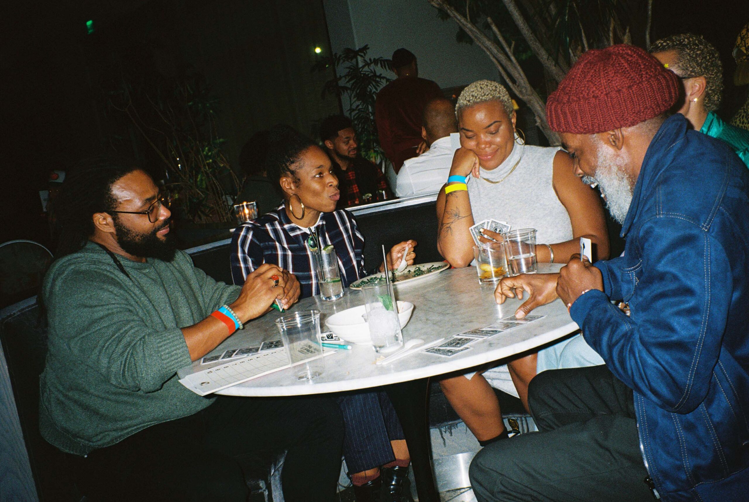 Five individuals at a table play cards, relishing their drinks in merriment.