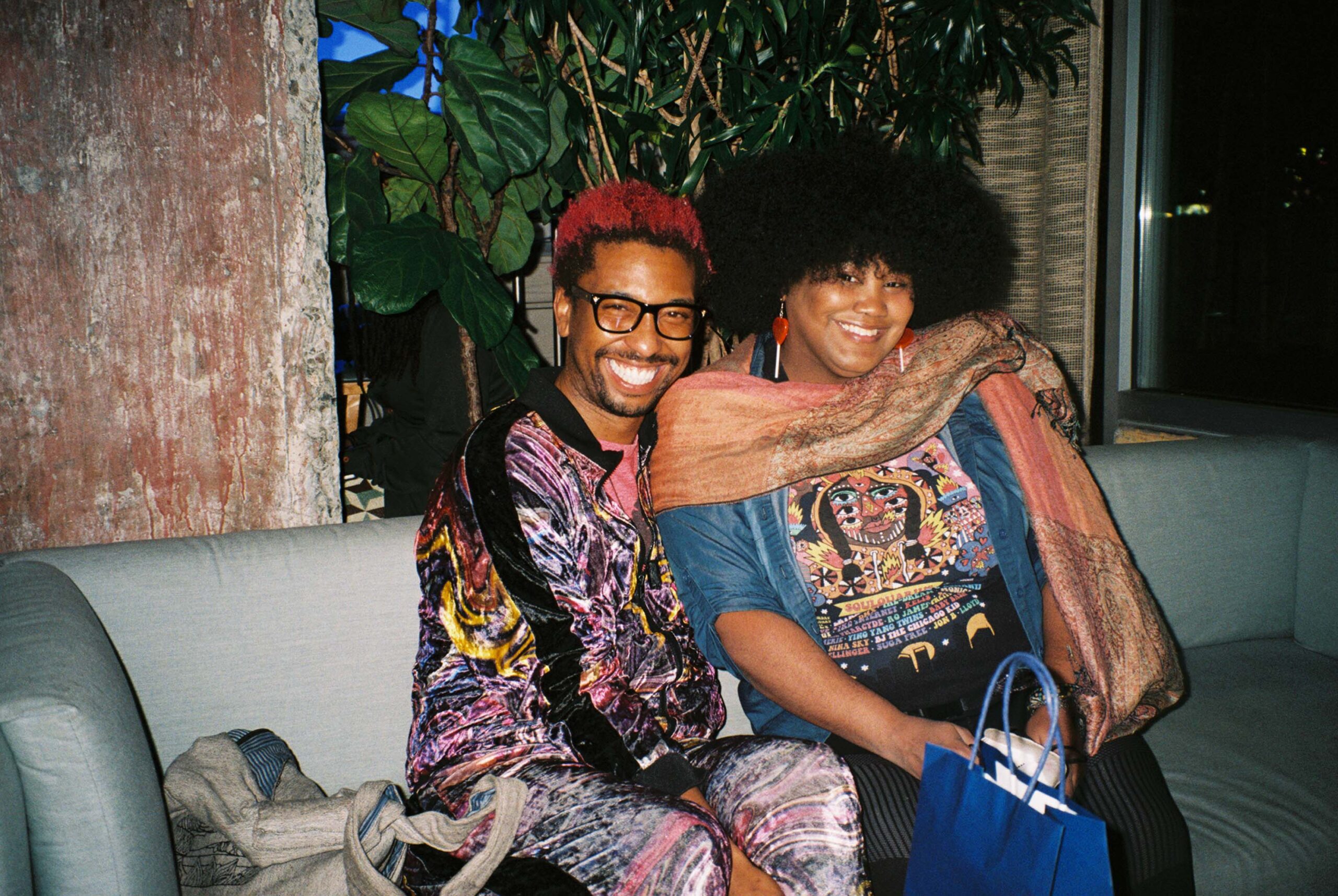 Two people sit on a sofa and pose in colorful outfits