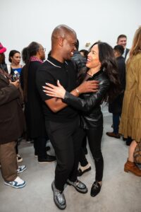 Two people excitedly greet and embrace at a busy art gallery opening