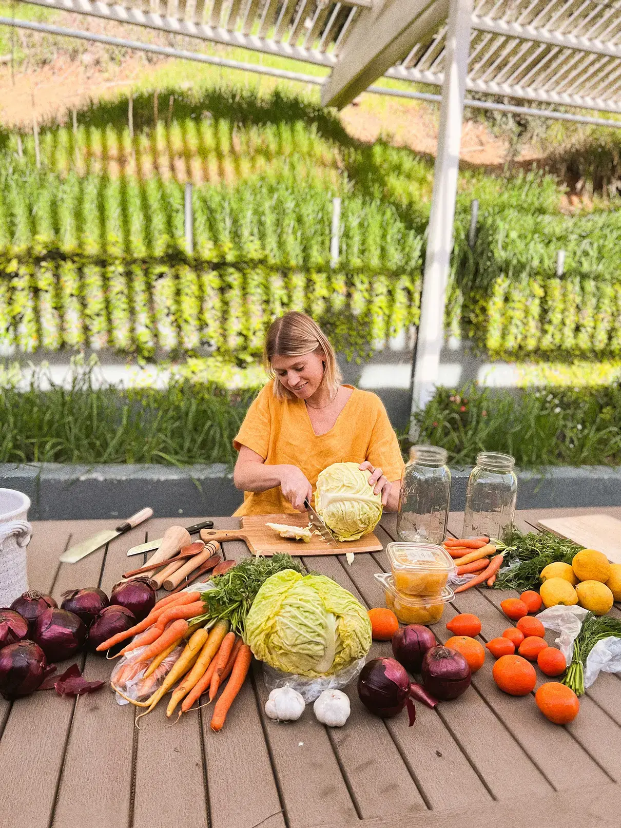 A woman surrounded by vegetables and mason jars is cutting a head of cabbage.