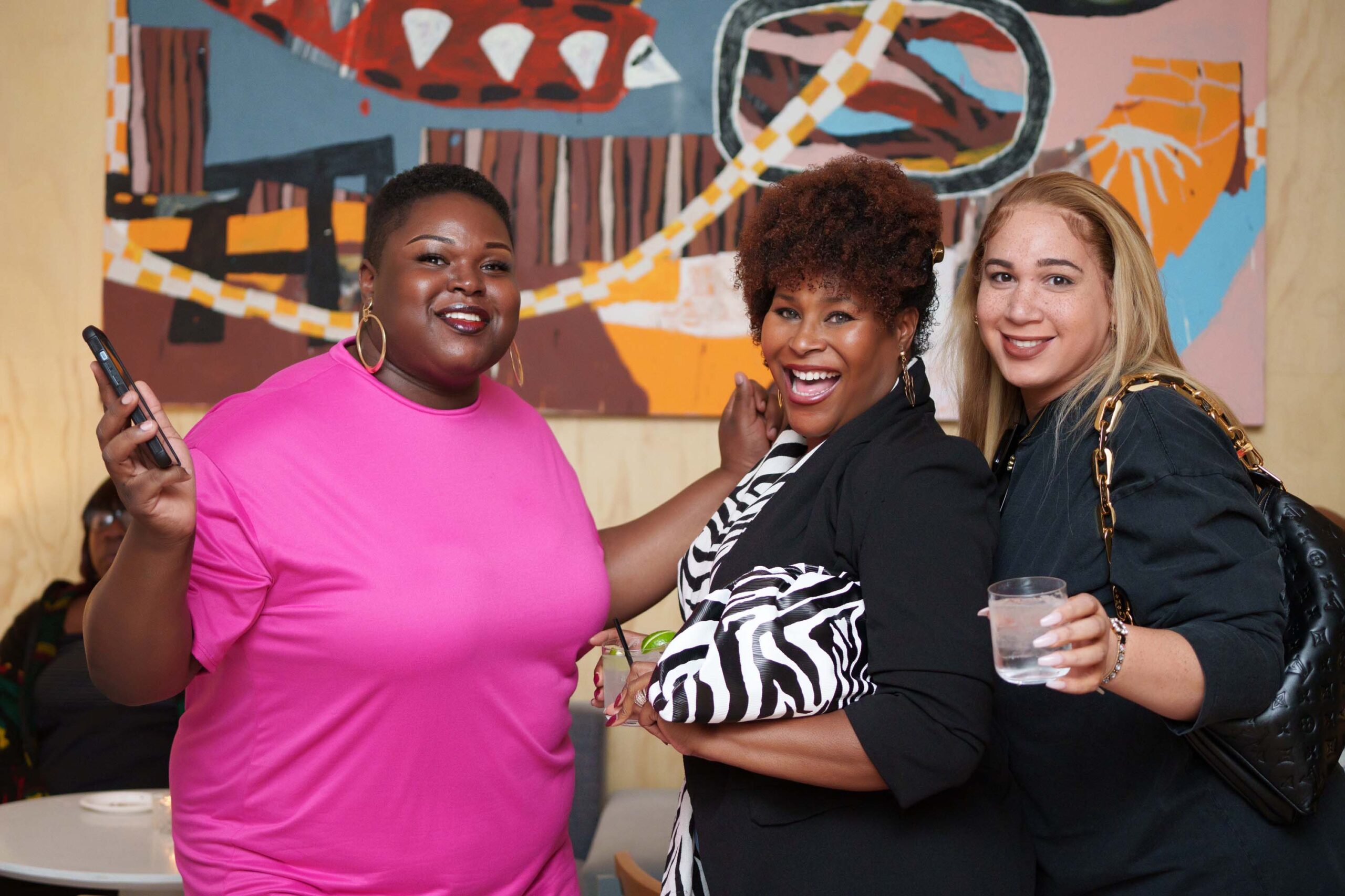 Three women smile happily in front of a large colorful painting