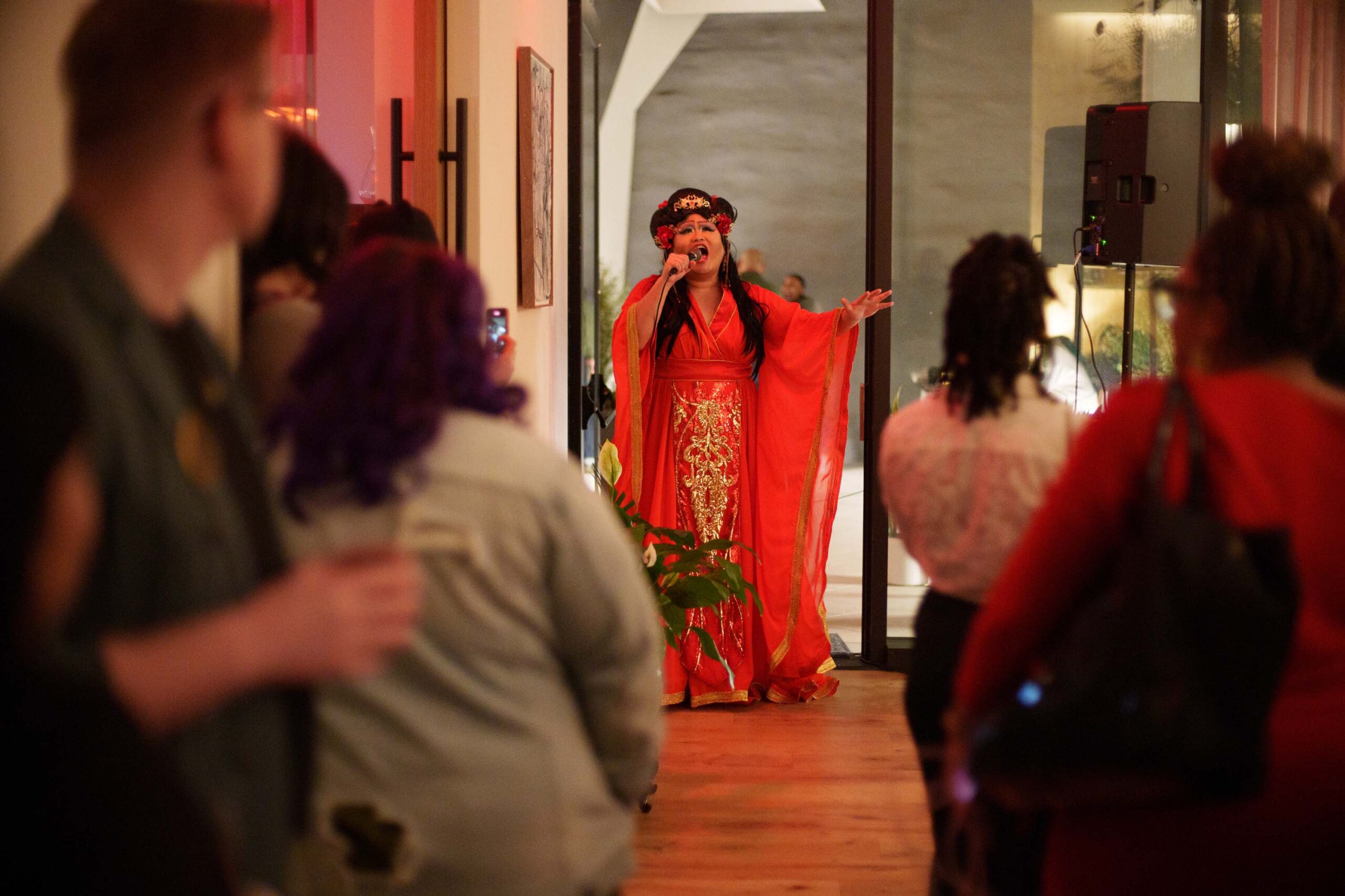 A drag queen in a red, traditional Chinese dress sings while people watch