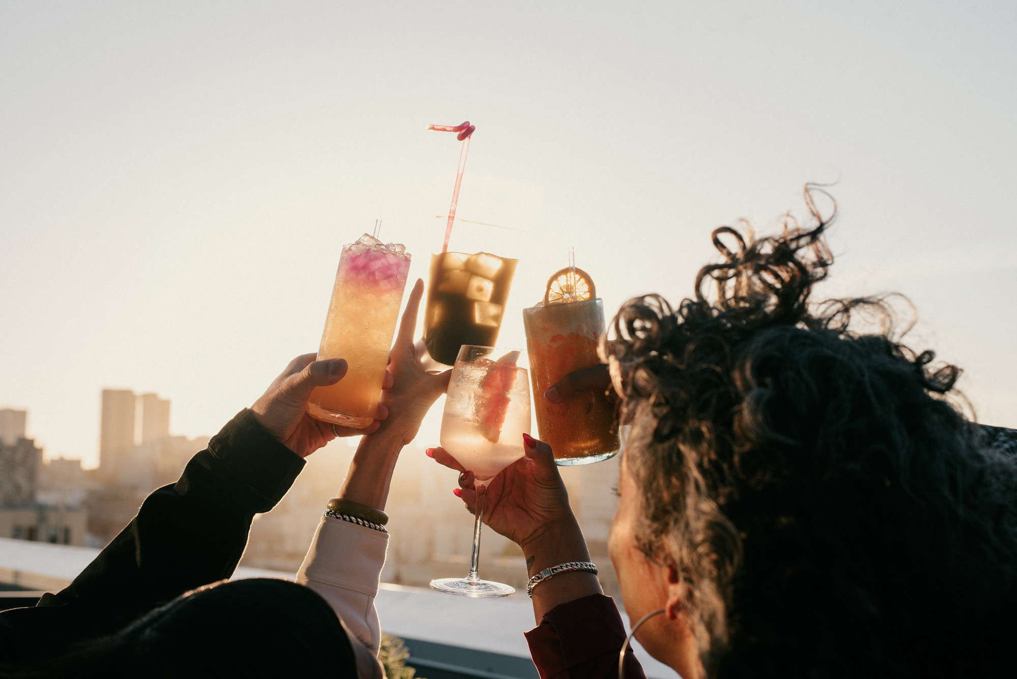 People's hands cheers with drinks above a city sunset skyline