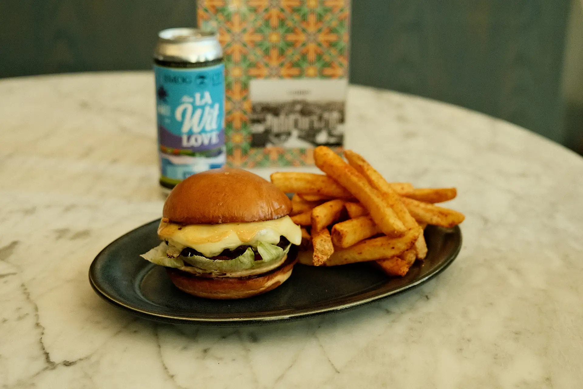 Cheeseburger with lettuce, fries, and canned drink on black plate atop a marble table.