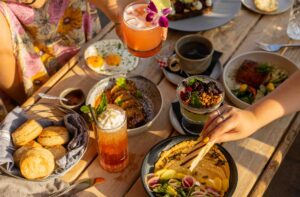 Sunny outdoor brunch table with colorful cocktails, biscuits, hummus, eggs, and shared small plates.