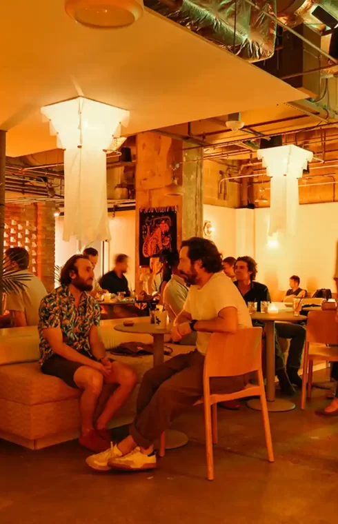 Dimly lit, modern lounge bar filled with people socializing, potted plants, warm lights and exposed ceilings.