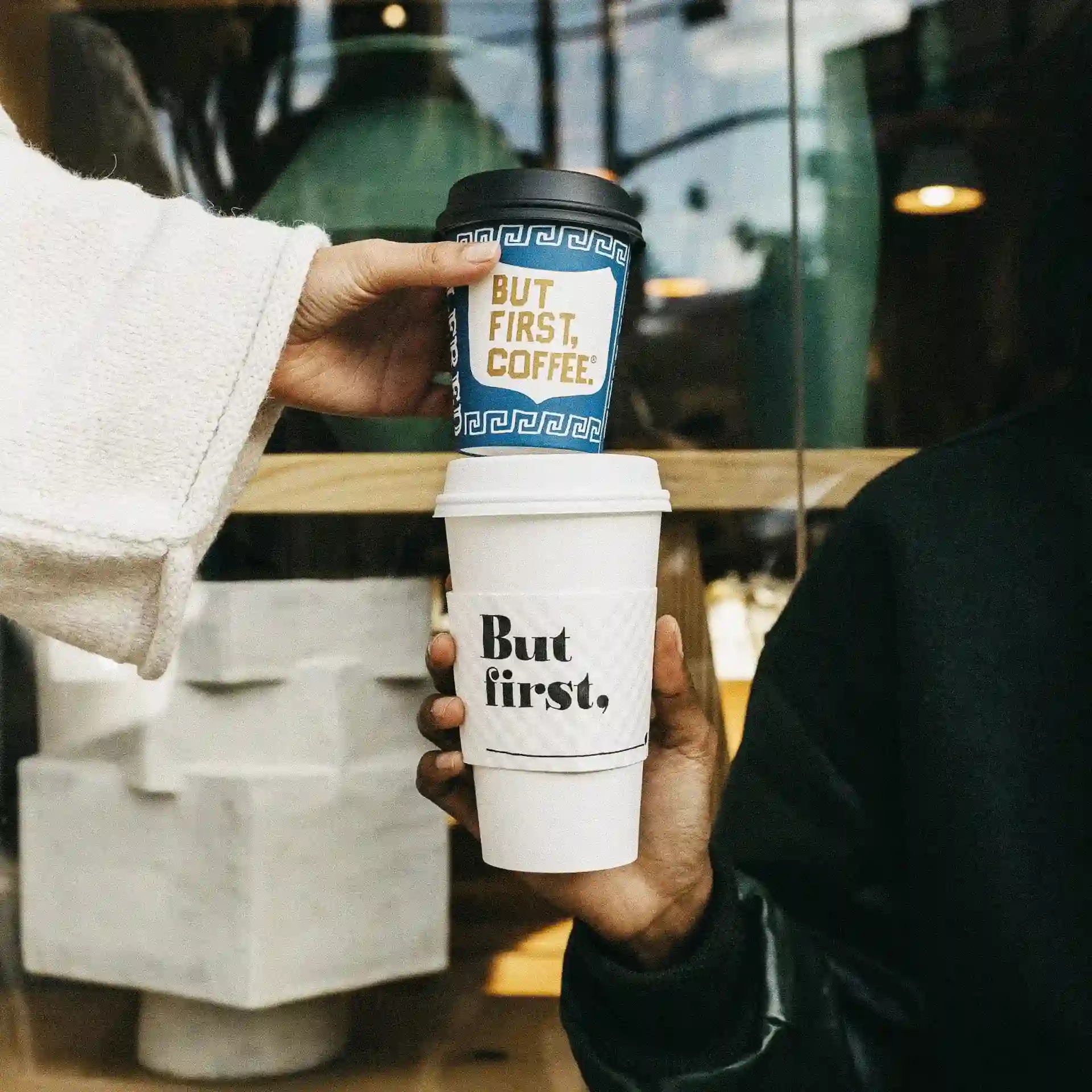 Two hands holding stacked coffee cups reading ‘But first, coffee’ outside a café window.