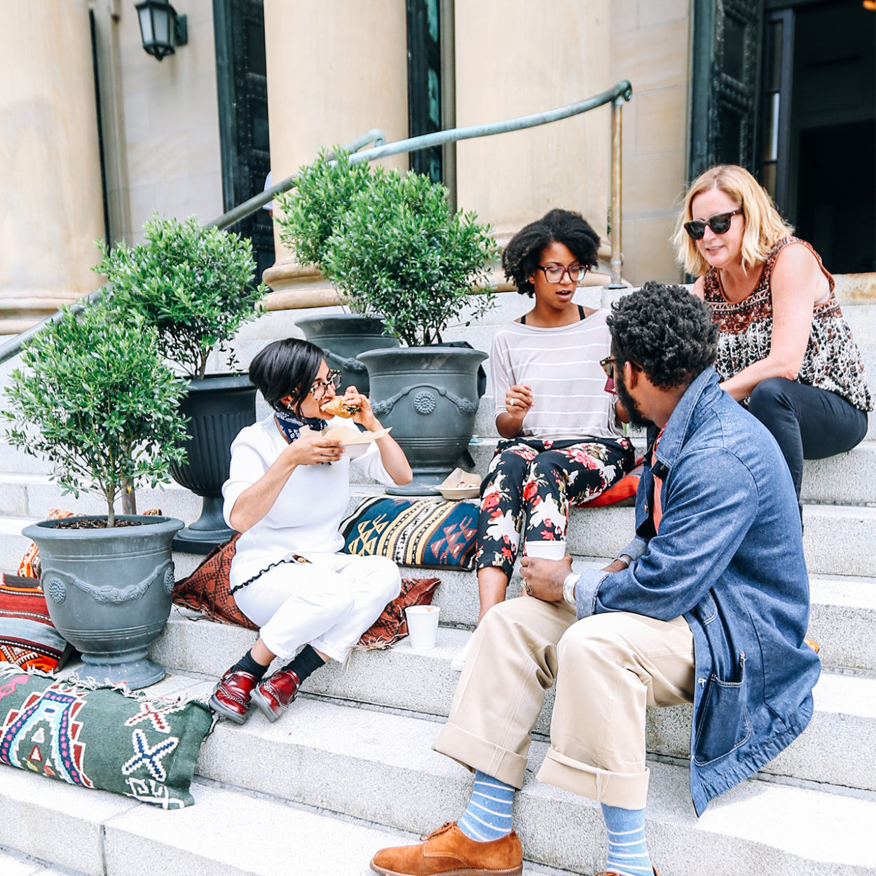 Four friends sit on outdoor steps with plants, eating and chatting in a relaxed atmosphere.
