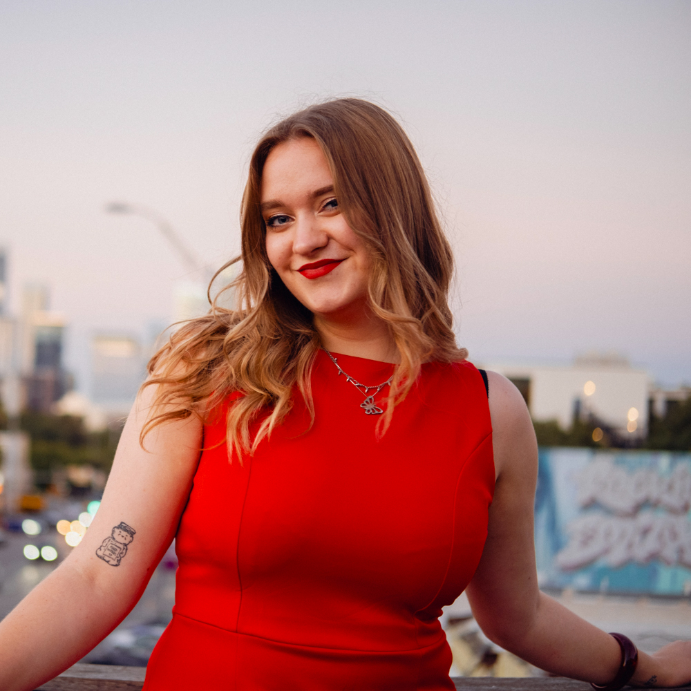 Smiling woman in a red dress standing outdoors at sunset, with a softly blurred city skyline behind her.