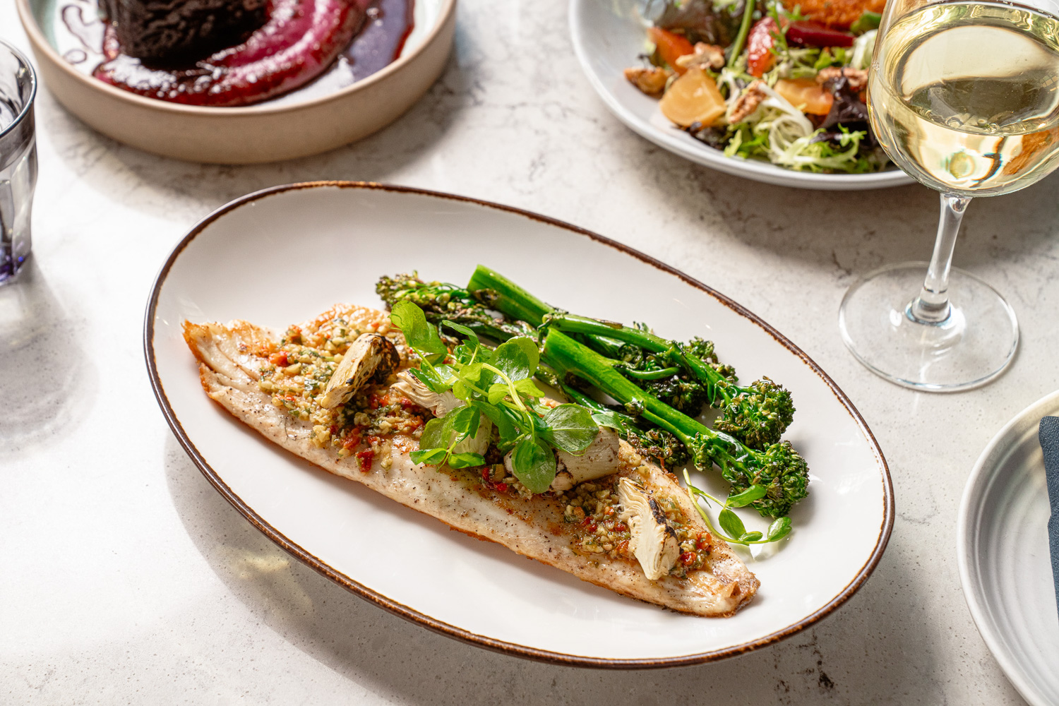 Seared fish fillet topped with herbs and mushrooms, served with broccolini on a white plate, with wine and salad nearby.