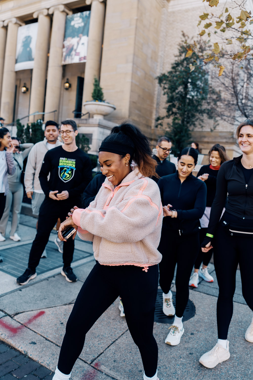 A group in athletic wear walks together outdoors, smiling as one person checks a smartwatch on city steps.