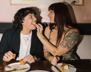 Two women smiling at a table as one feeds the other a bite of food, plates and drinks in front of them.