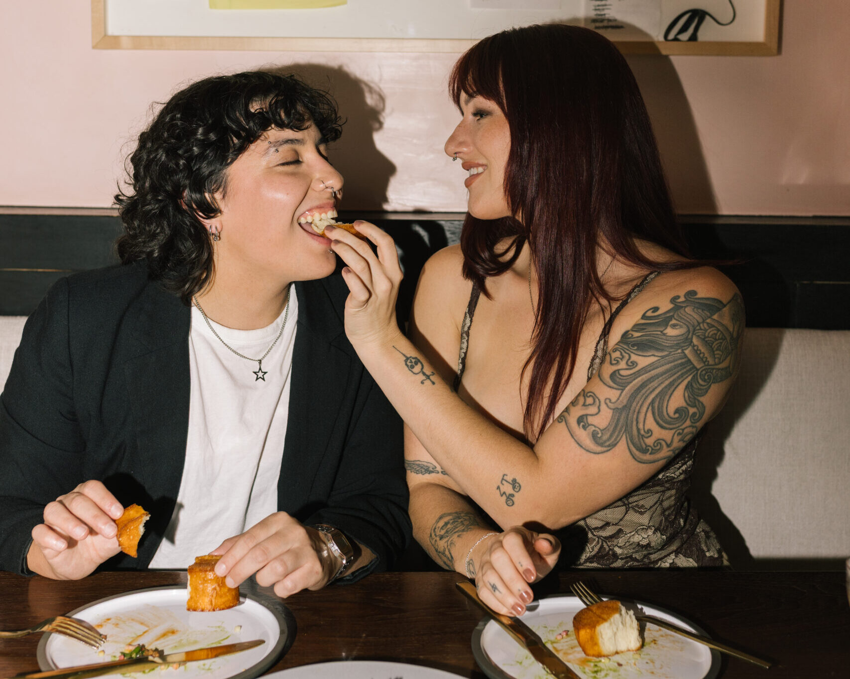 Two people laughing at a restaurant table as one feeds the other a bite of food, sharing a playful, intimate dining moment.