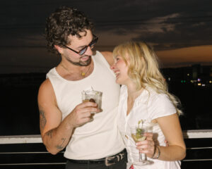 Smiling couple holding drinks on balcony at sunset with city lights in background.