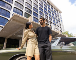 Couple standing by vintage car outside modern hotel building on sunny day