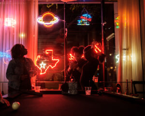 Silhouetted people playing pool in bar with colorful neon signs glowing in window.