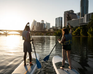 Two people paddleboarding on river at sunset with city skyline and bridge in background.