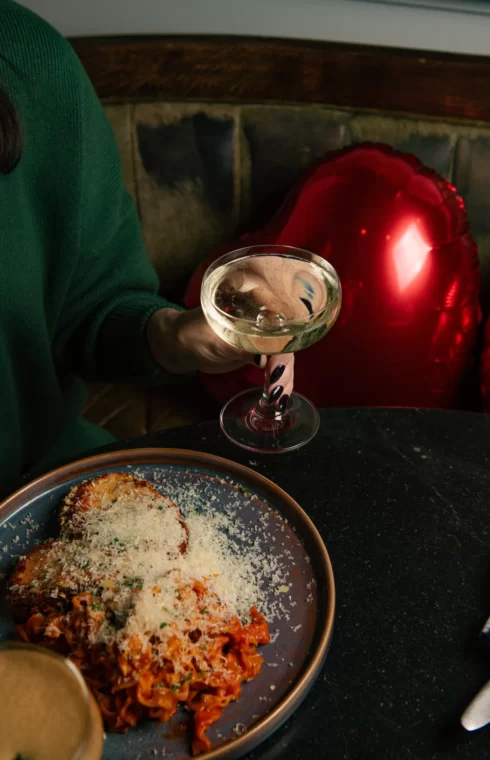 Person holding a coupe cocktail beside a plate of pasta with grated cheese, seated at a table with a red heart balloon.