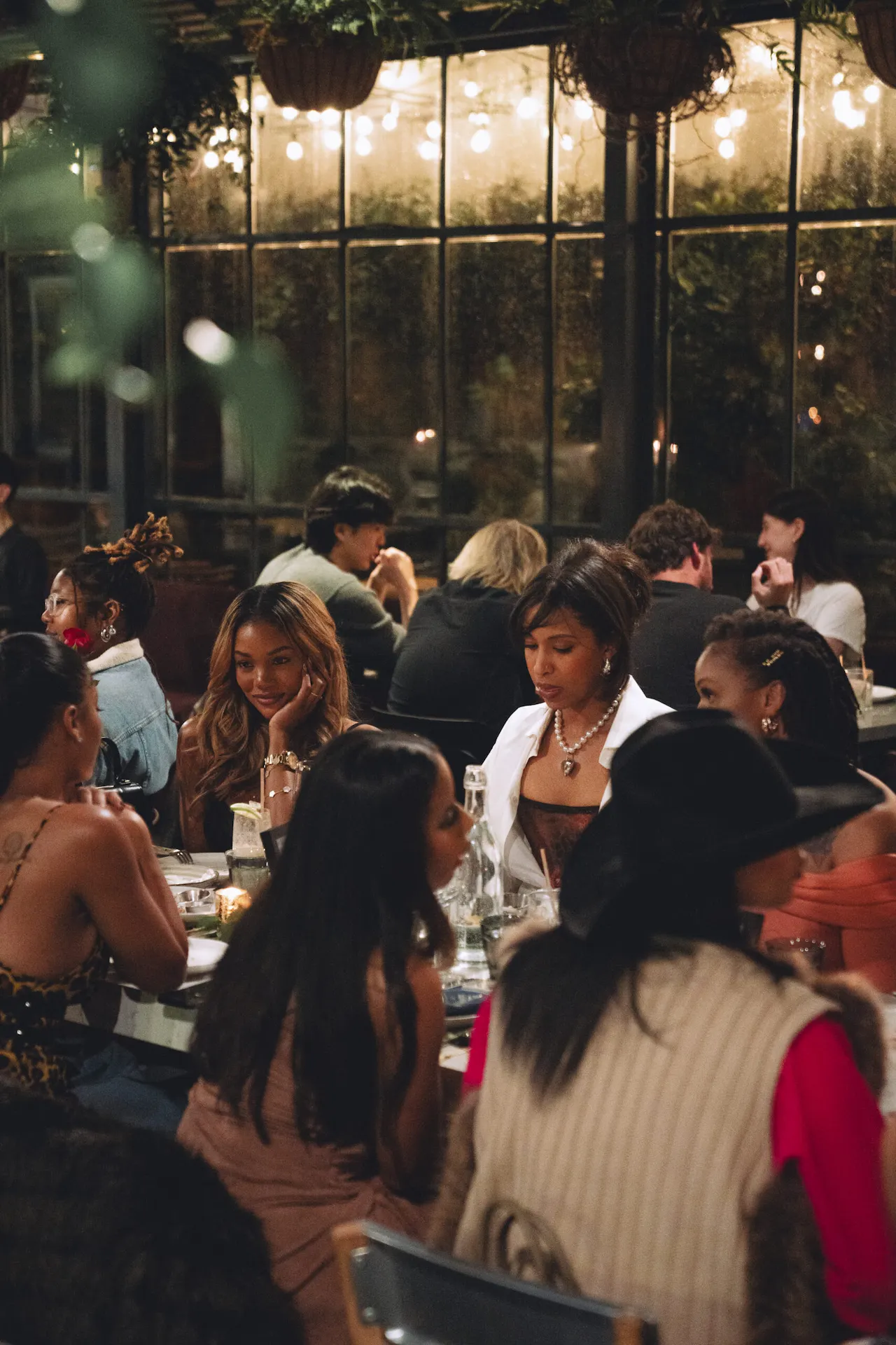 Group of women dining together at a warmly lit restaurant with hanging plants and lively conversation.