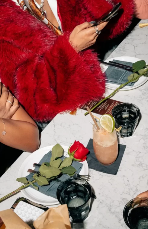 Hands at a restaurant table with cocktails, water glasses, and red roses; one person wears a red faux fur coat.
