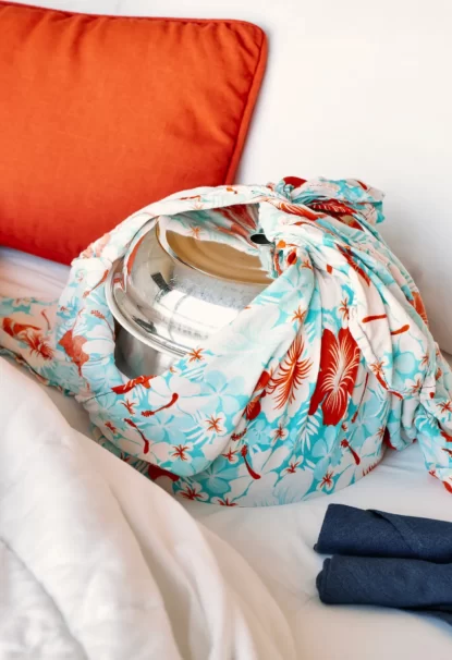 Shiny metal pot wrapped in colorful floral fabric, resting on a bed with an orange pillow and two rolled blue towels nearby.