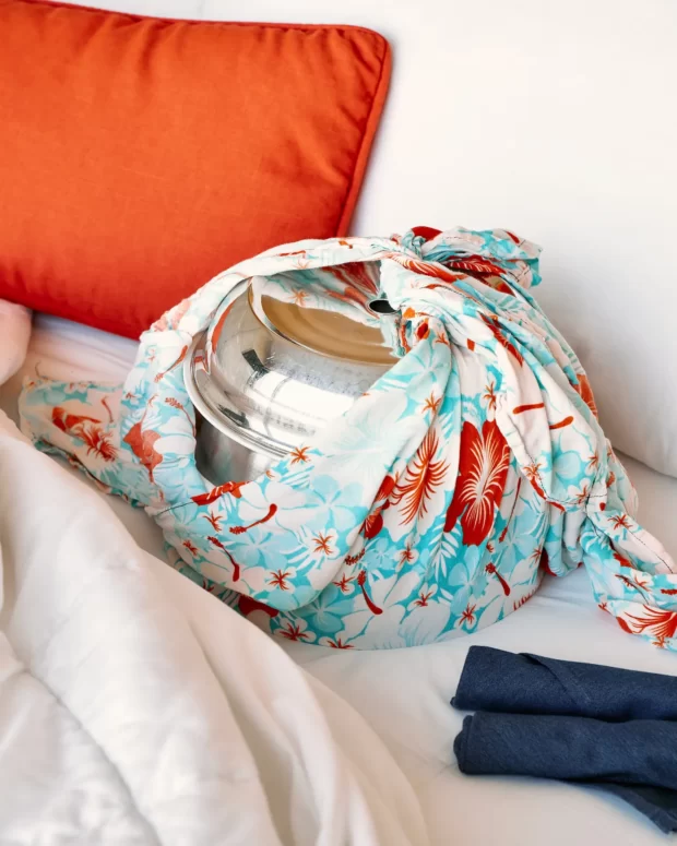 화려한 꽃무늬 천으로 감싸인 반짝이는 금속 냄비가 주황색 베개와 두 개의 접힌 파란색 수건이 놓인 침대 위에 놓여 있습니다.