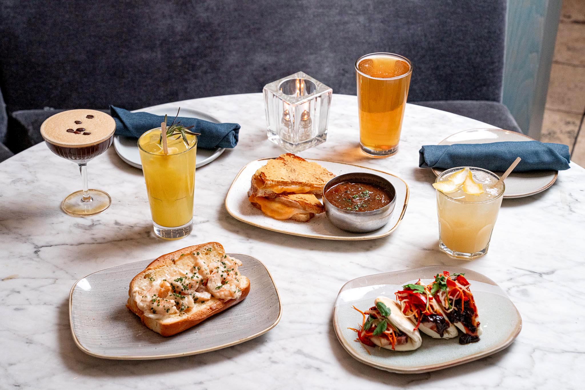 Marble table with assorted dishes, bao buns, sandwiches, cocktails, and beer arranged for a shared meal.