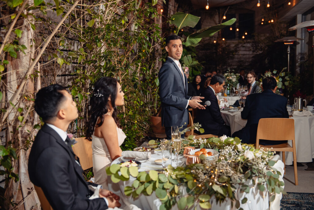 Married couple is sitting in the dining table with other people and a anchor is standing looking at couple