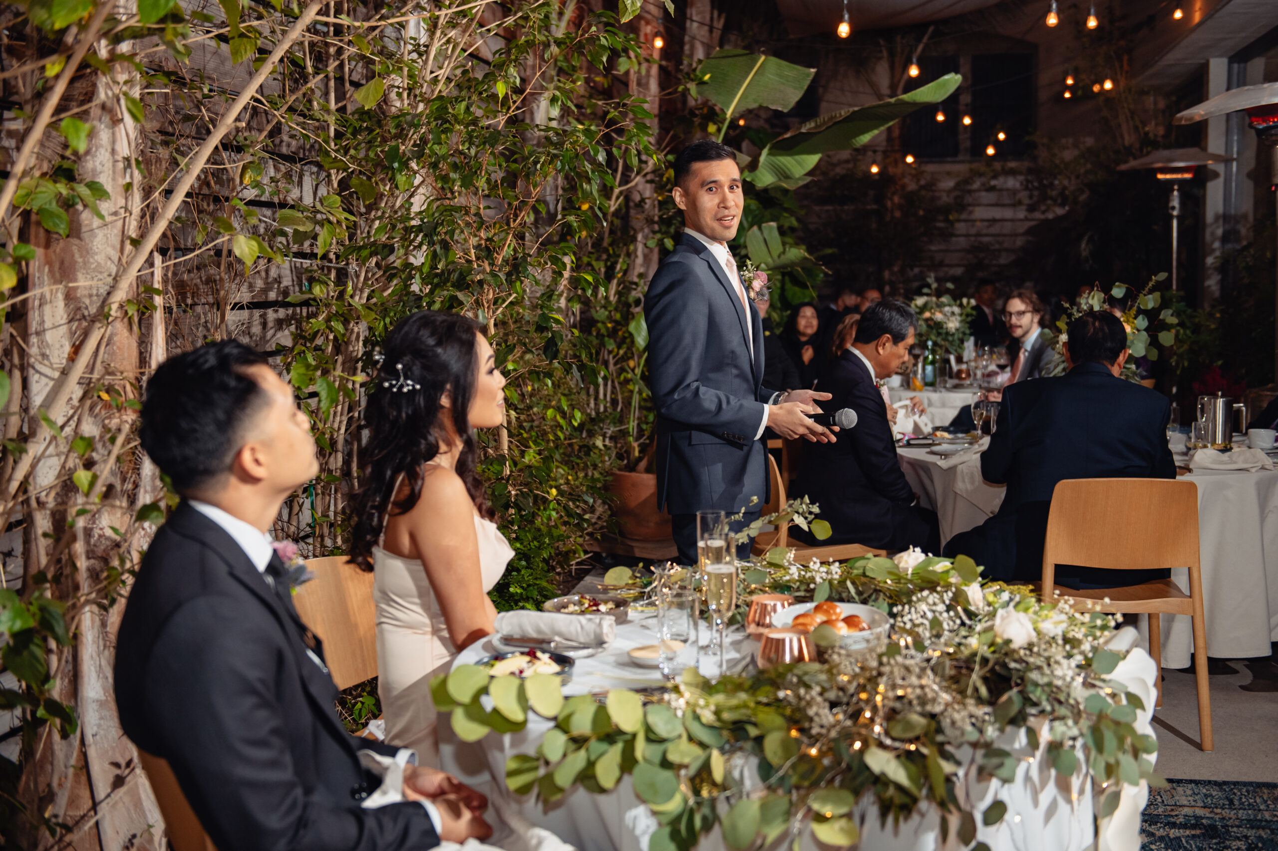 Married couple is sitting in the dining table with other people and a anchor is standing looking at couple