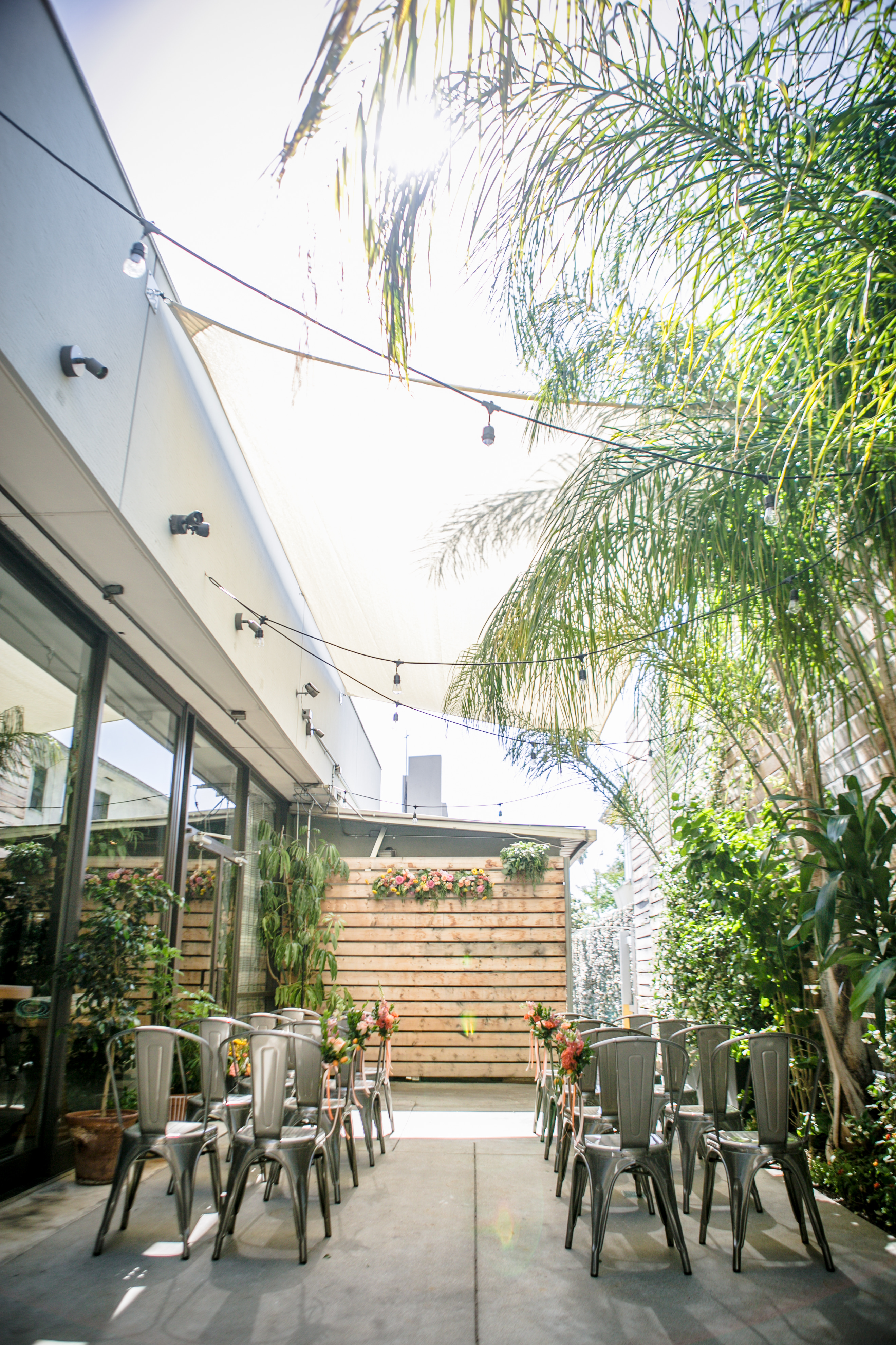 A serene outdoor space with metal chairs arranged for an event, surrounded by palm trees and flowers under string lights.