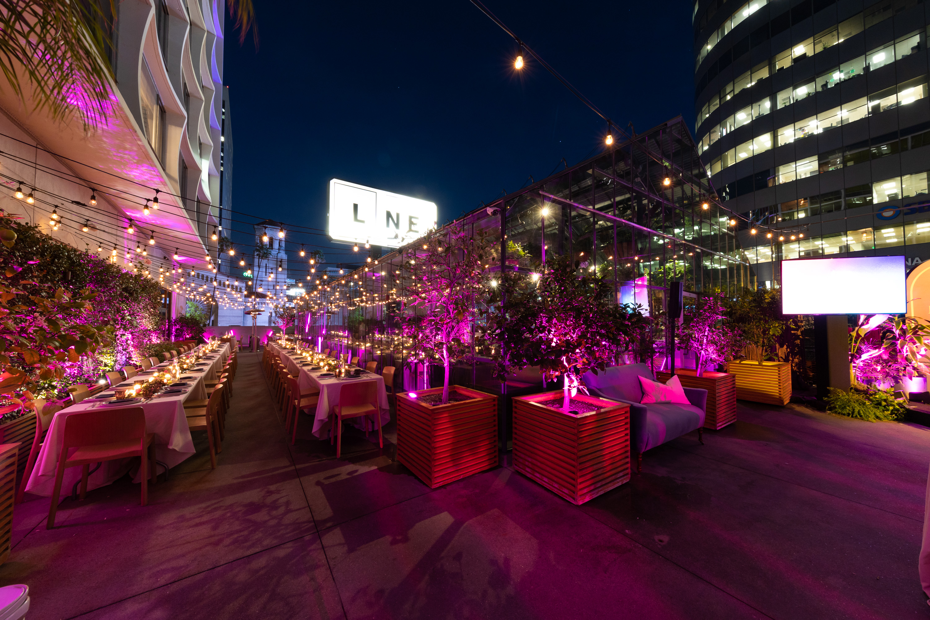 A rooftop event space at night with tables, chairs, string lights, and decorative plants.