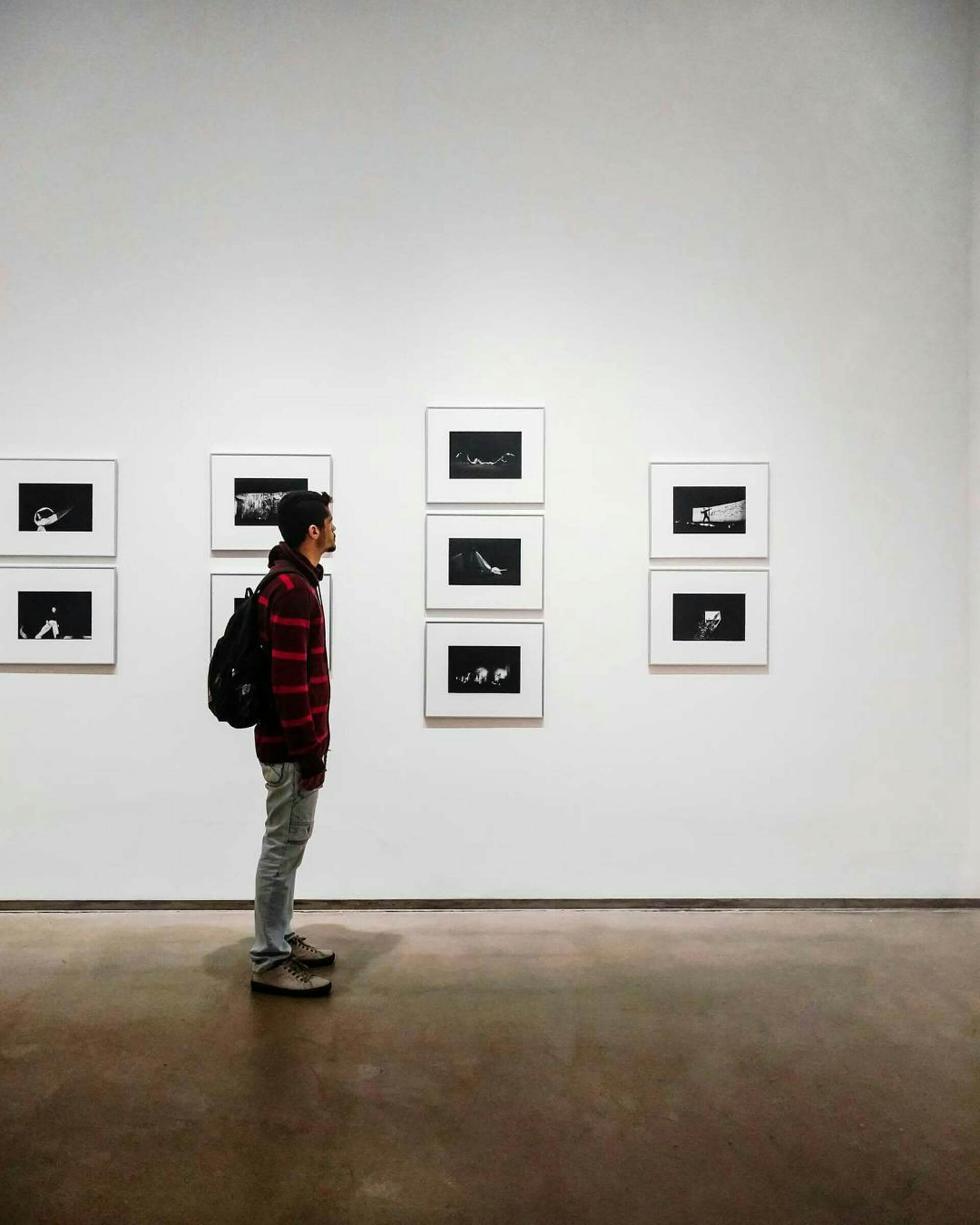 Man with a backpack viewing framed black-and-white photos in an art gallery.