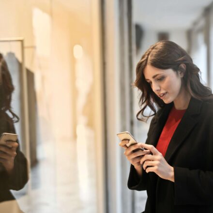Woman using a smartphone while window shopping, with her reflection visible on the glass.