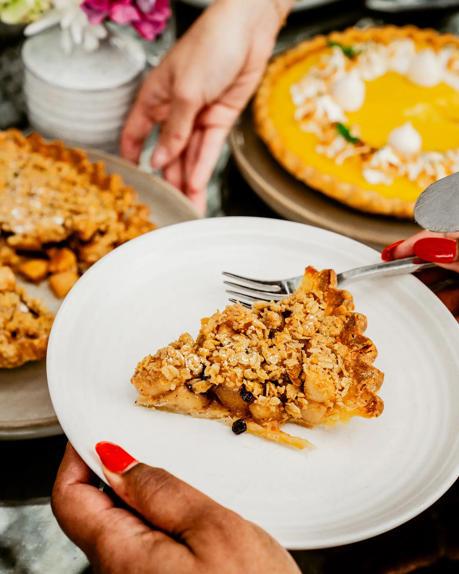 Slice of apple crumble pie being served onto white plate, with hand and festive dessert table in background