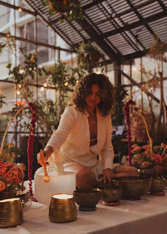 Thalia Ayres Randolph playing crystal singing bowl in indoor garden setting with candles, flowers, and string lights