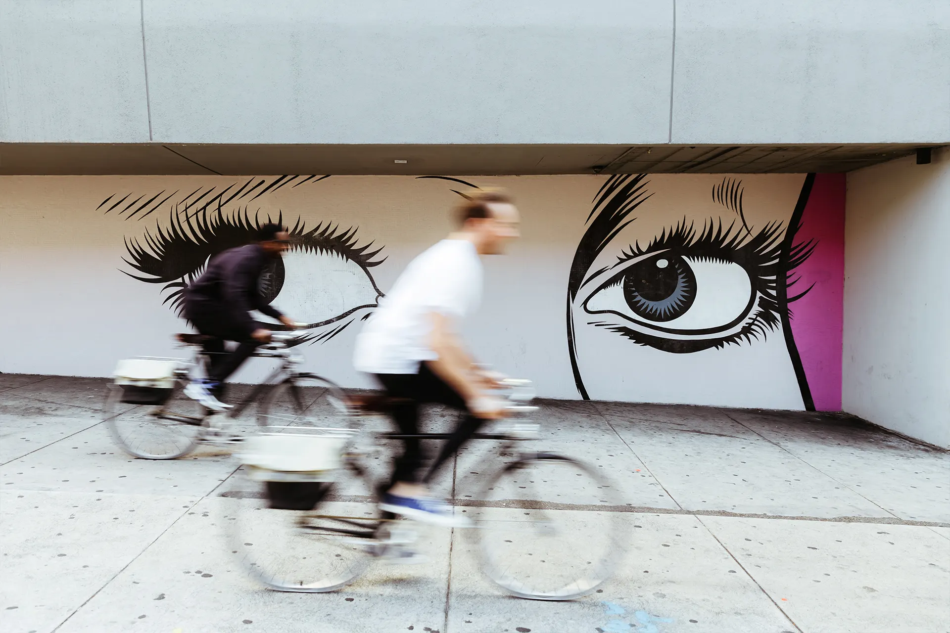 Two cyclists passing mural of stylized eyes with motion blur on concrete wall under overhang
