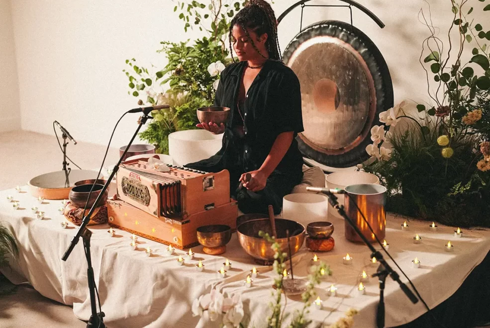 Person seated with singing bowl amid candles, plants, and sound healing instruments in serene indoor ritual setting