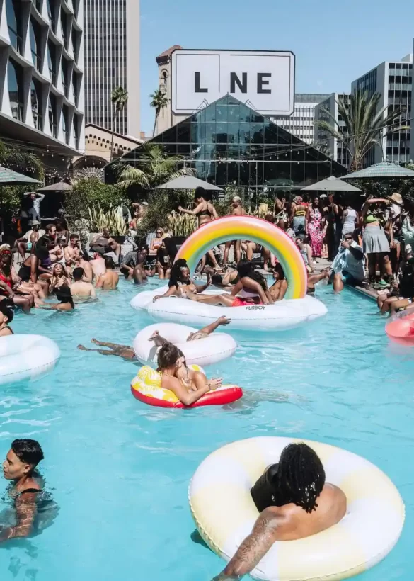 Crowded pool party at The LINE LA hotel with colorful floats, palm trees, and city buildings in the background