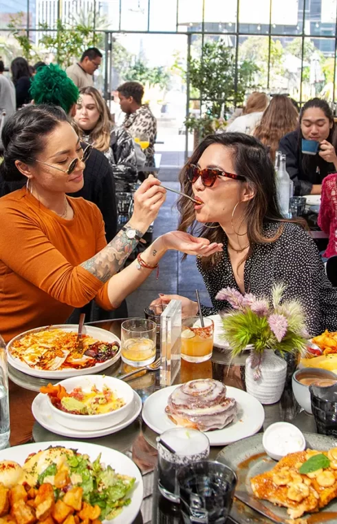 Two people sharing food at Openaire restaurant table filled with colorful dishes, greenery, and natural light